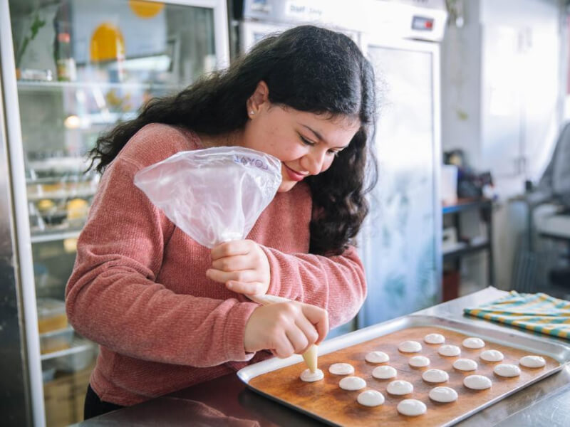 A smiling woman pipes macaron shells onto a baking sheet with a plastic piping bag